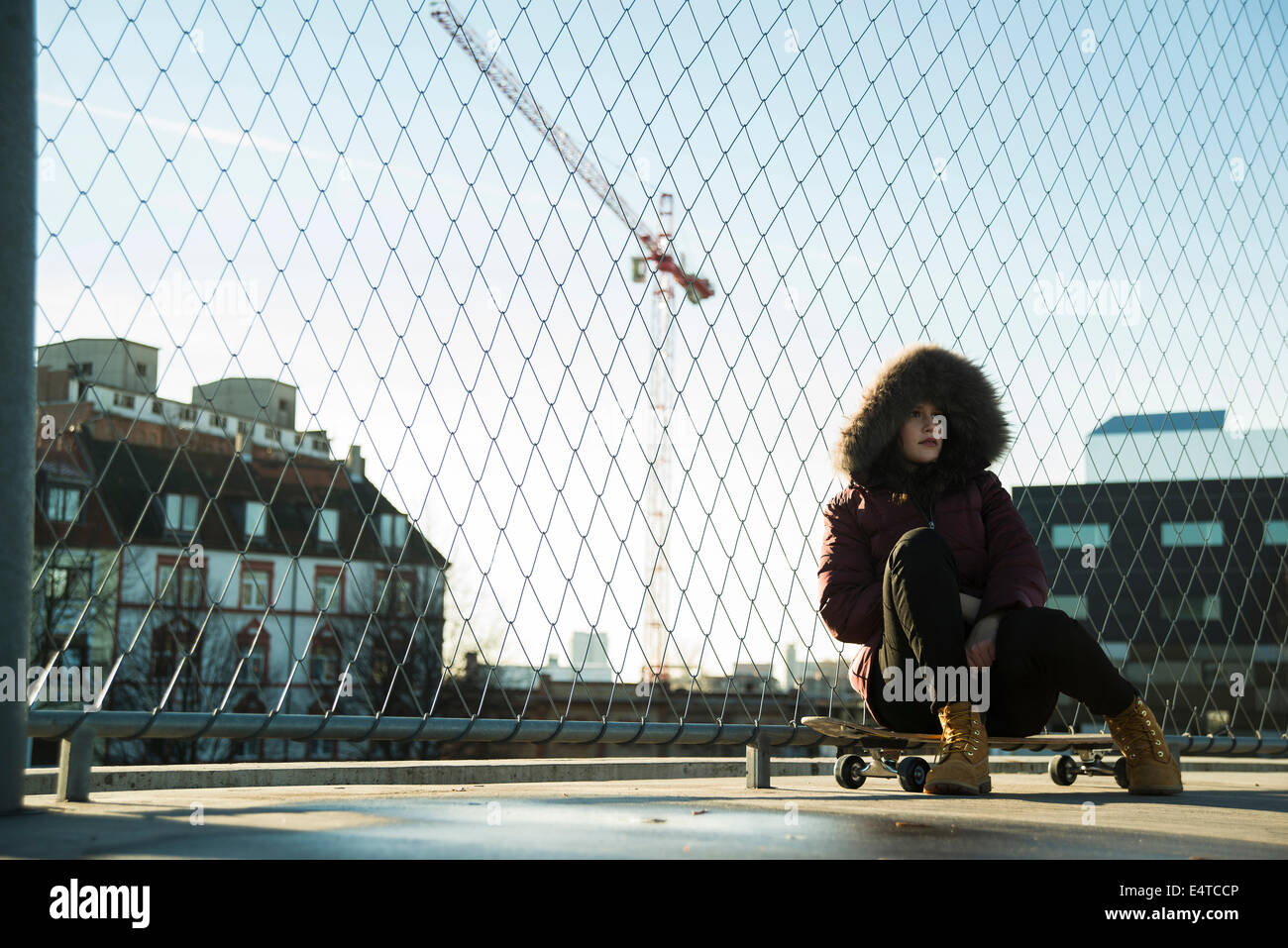 Teenage girl wearing winter coat, sitting on skateboard outdoors, next to chain link fence near comercial dock, Germany Stock Photo