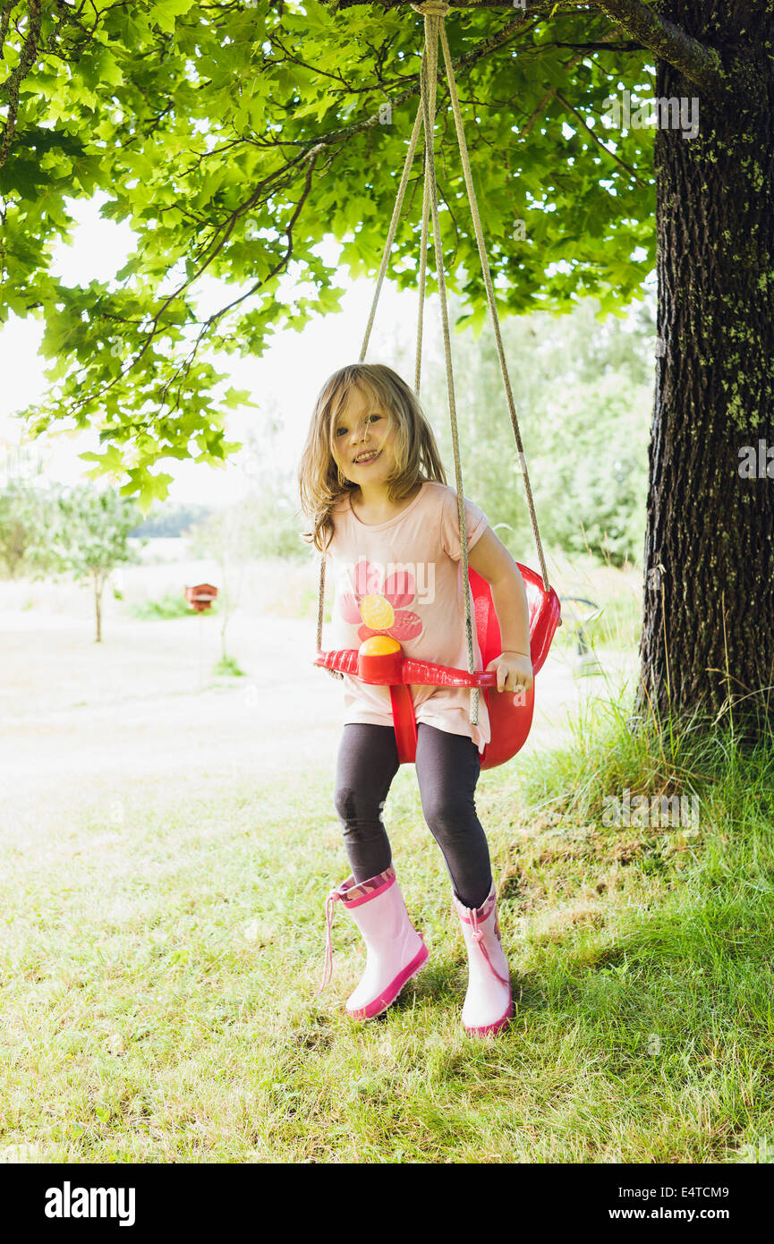 3 year old girl in rubber boots sitting in red swing in back yard, looking at camera and smiling, Sweden Stock Photo
