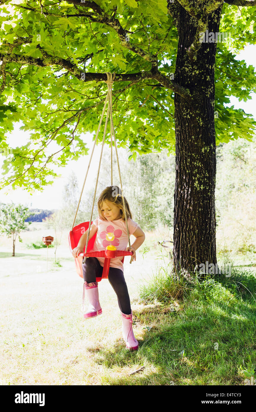 3 year old girl in rubber boots sitting in red swing in back yard, Sweden Stock Photo