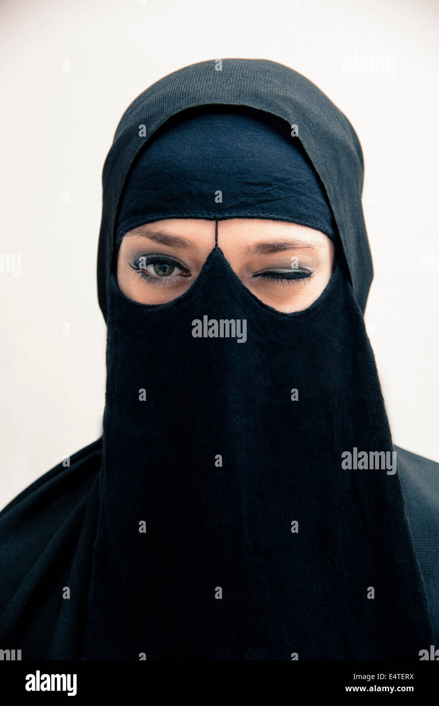 Portrait of young woman in black, muslim hijab and muslim dress, winking at camera, eyes with makeup, on white background Stock Photo