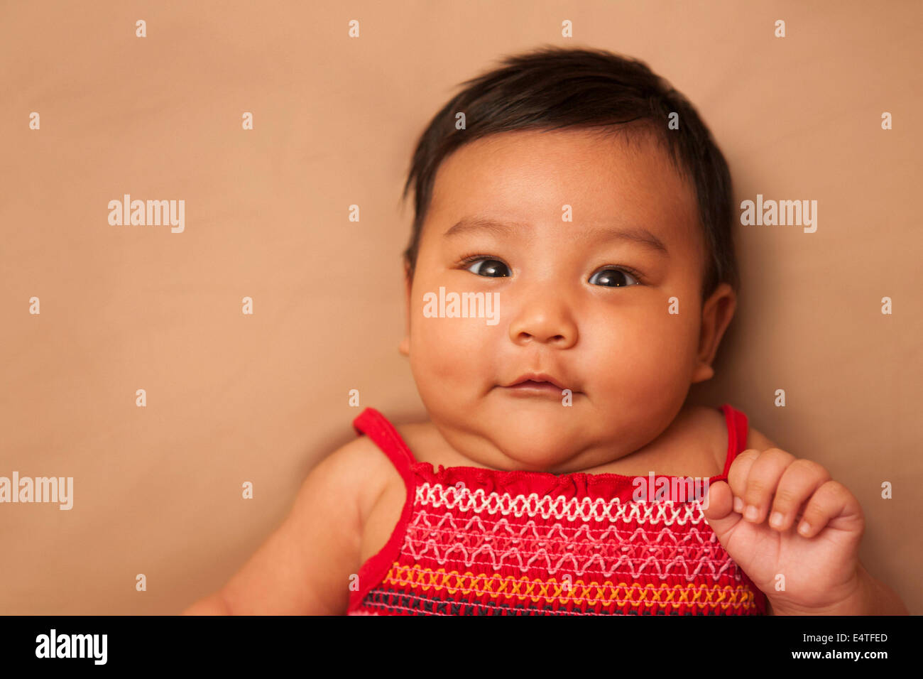 Close-up portrait of Asian baby lying on back, wearing red dress, looking at camera and smiling, studio shot on brown background Stock Photo