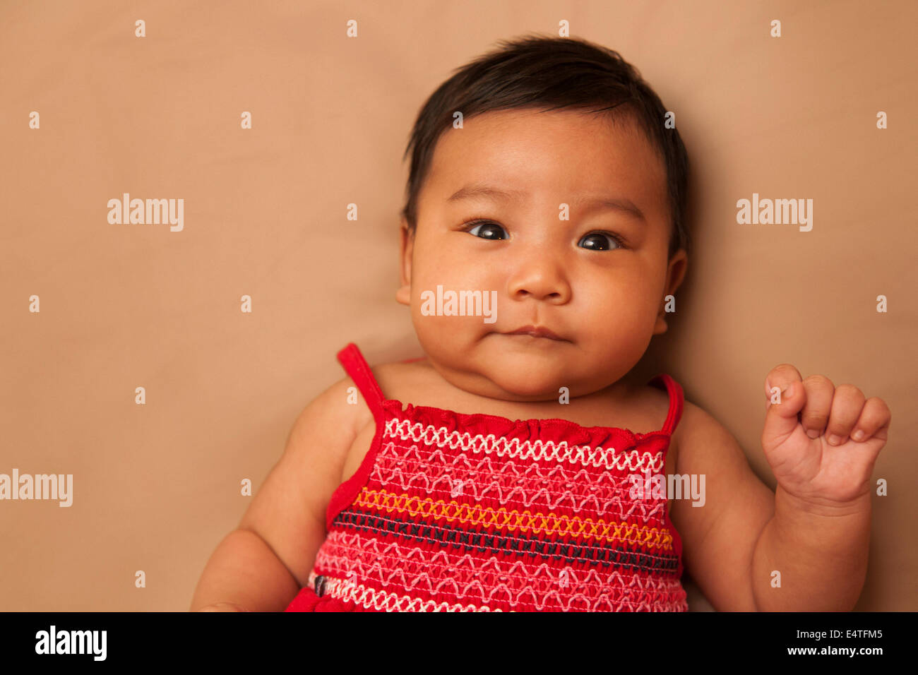 Close-up portrait of Asian baby lying on back, wearing red dress, looking at camera and smiling, studio shot on brown background Stock Photo