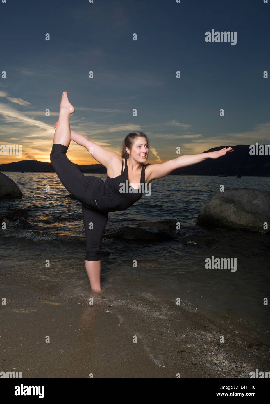 An attractive young woman in black spandex strikes a yoga pose in the waves of a beach at sunset. Stock Photo