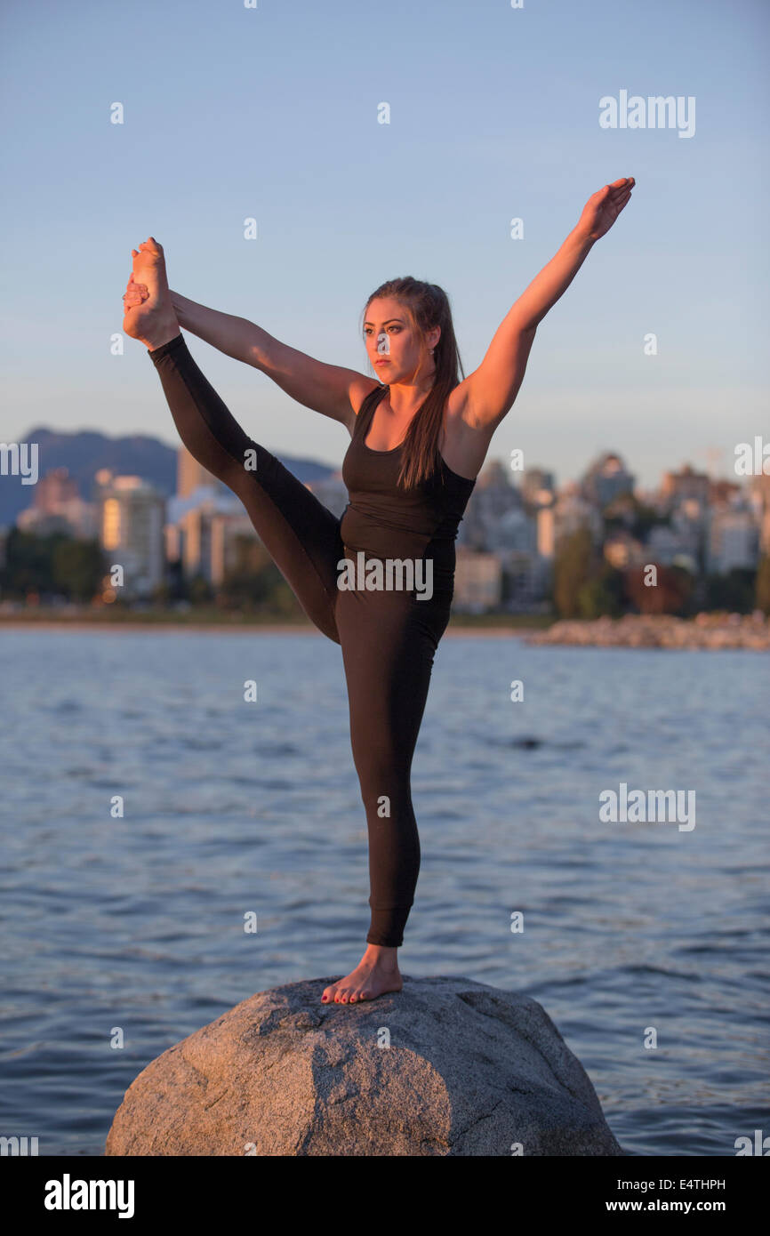 An attractive young woman in fitness wear stands on a rock on one leg and stretches the other leg with the ocean and downtown Va Stock Photo