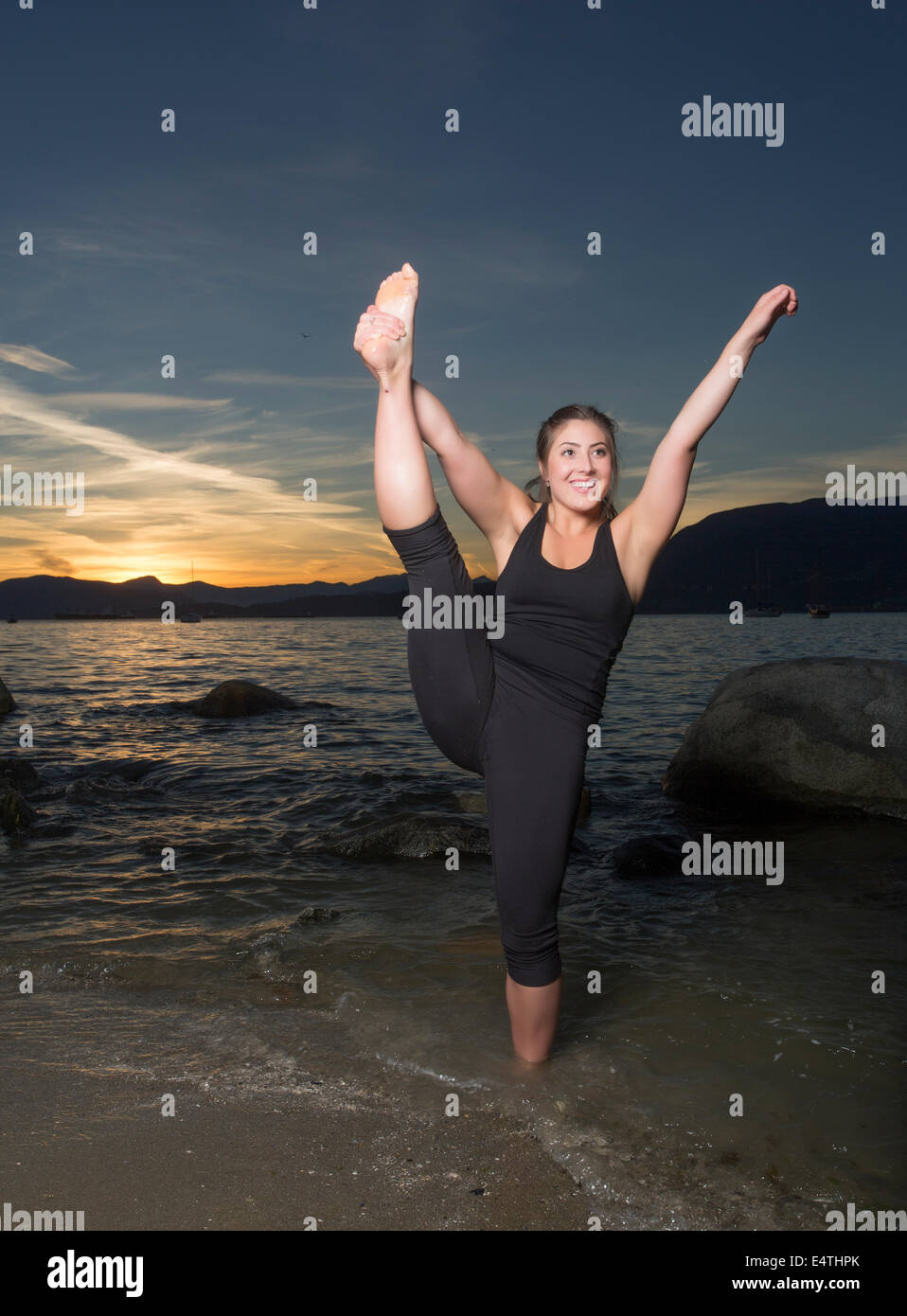 An attractive young woman stands one leg and stretches on the beach at sunset. Stock Photo