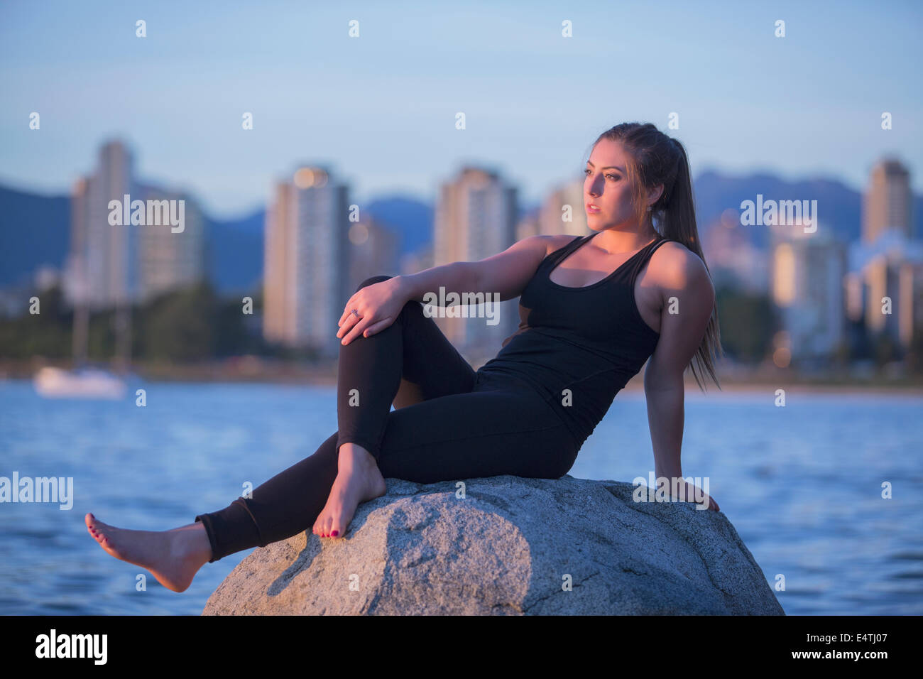 An attractive young woman relaxes on a rock with the ocean and the buildings of Vancouver behind her. Stock Photo