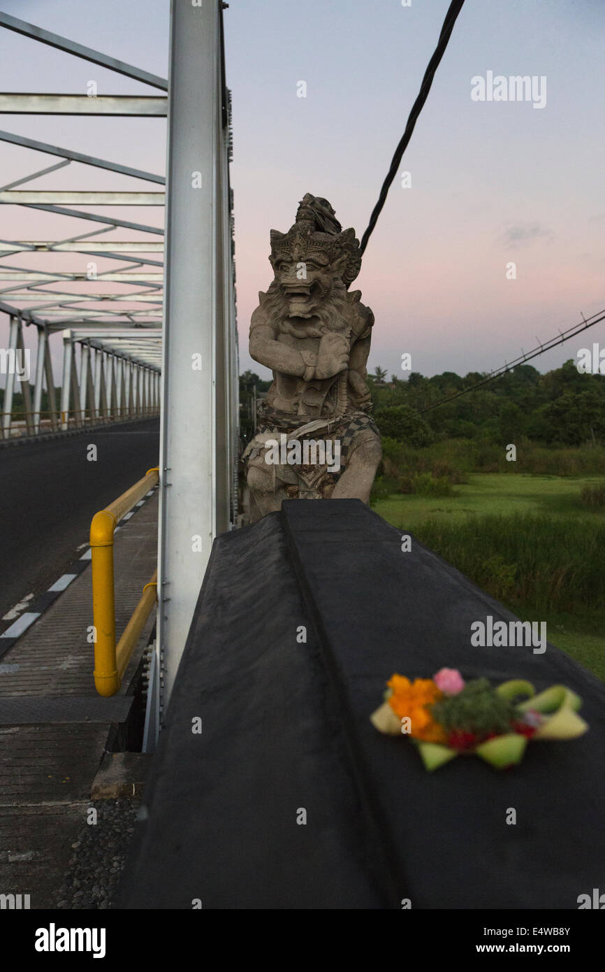 Bali, Indonesia.  Mythical Hindu Figure Guards a Bridge in Southern Bali.  Offering Basket (Canang) in Foreground. Stock Photo