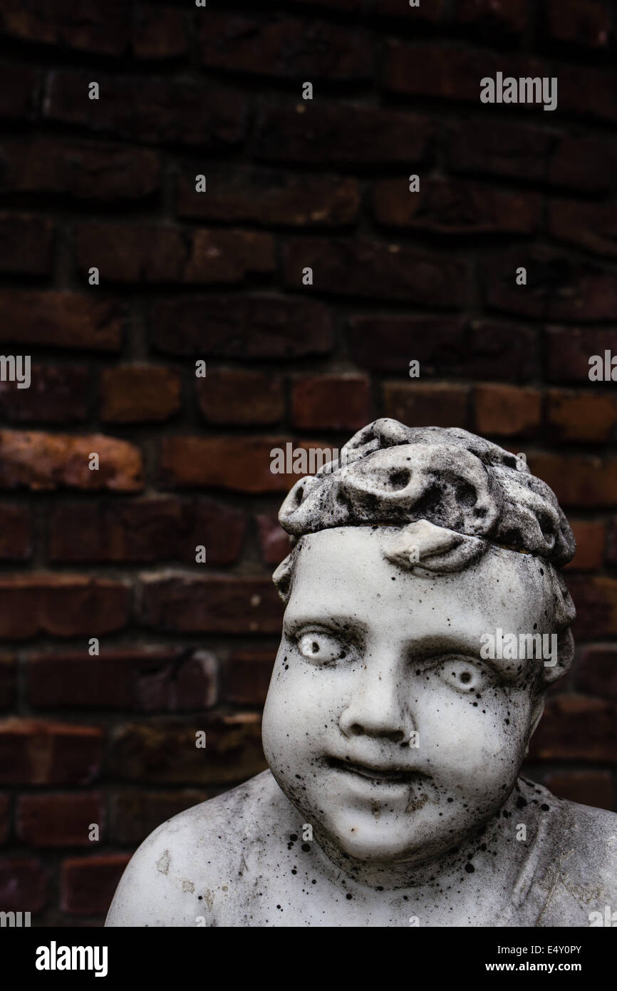 An old creepy vintage marble garden statue of a cherub or boy against and old brick wall.  Copy space above. Stock Photo