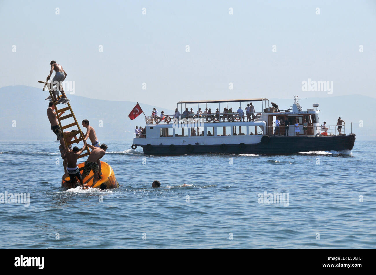 Turkish youths play on a buoy off the shore of Kinaliada, one of the Princes' islands, while a little ferry carrying day-tripper Stock Photo