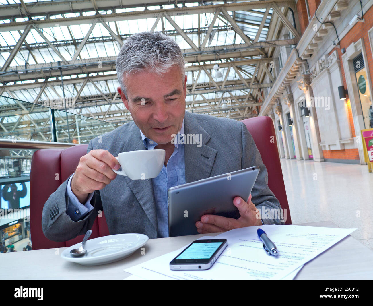 Mature businessman seated at cafe table on railway concourse looking at his tablet computer with iPhone and notes in foreground Stock Photo