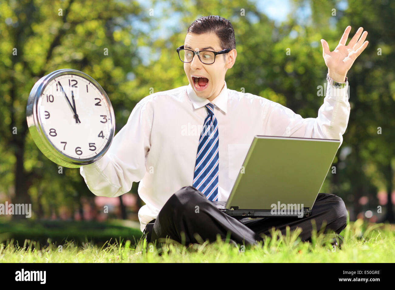 Young angry businessperson with computer sitting on grass and looking at clock in a parK Stock Photo