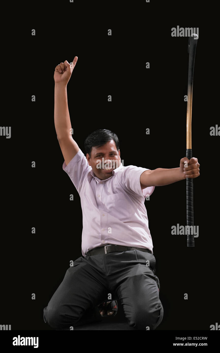 Portrait of happy Indian businessman with hockey stick pointing upwards while celebrating victory isolated over black background Stock Photo