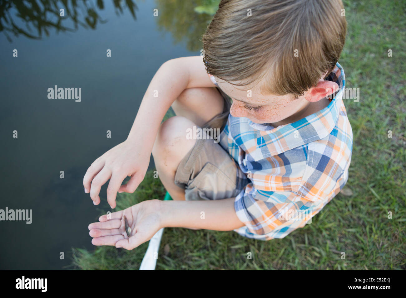 A young boy outdoors sitting on a riverbank with a small fish in the palm of his hand. Stock Photo