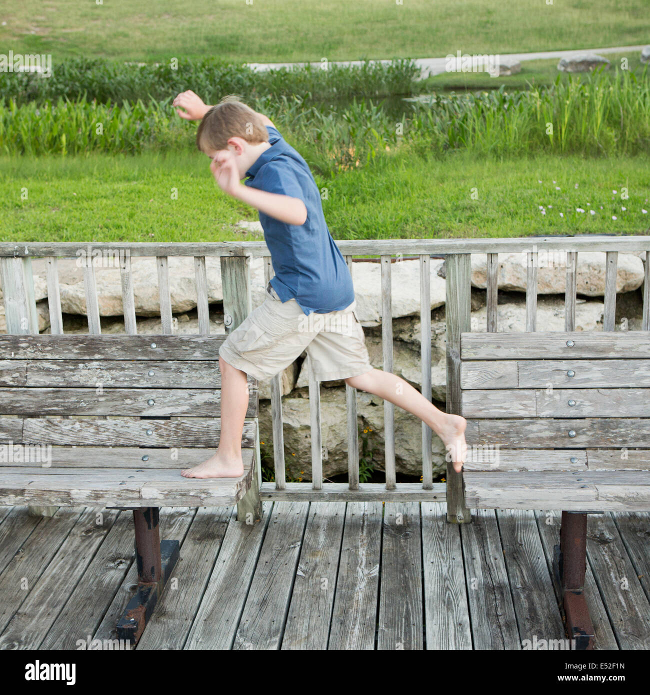A young boy outdoors leaping from one bench to another on a jetty over water. Stock Photo