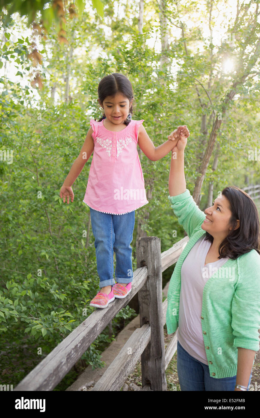 A young girl in a pink shirt and jeans, walking along a fence holding her mother's hand. Stock Photo