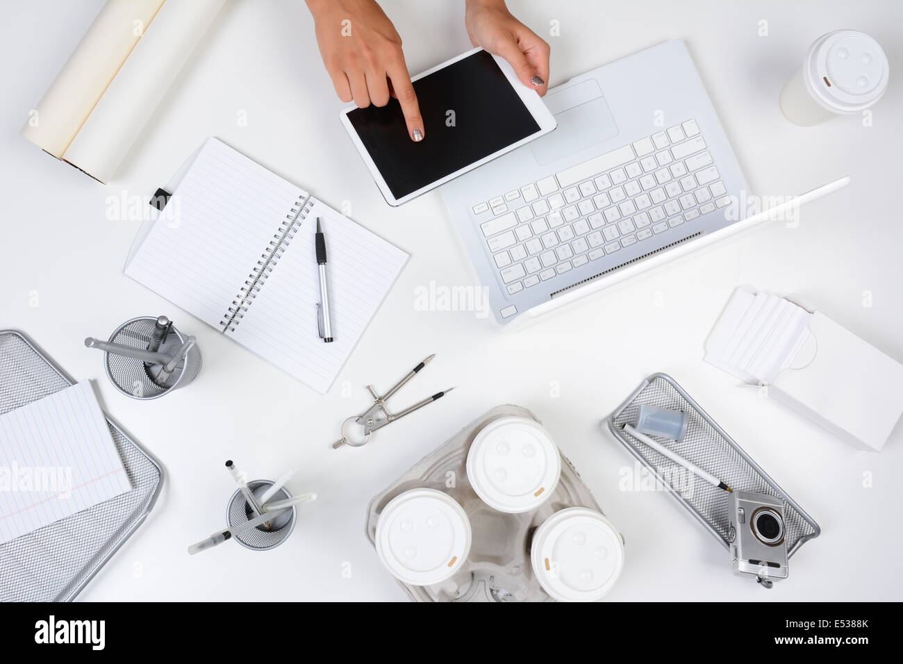 High angle shot of a white desk with primarily white and silver office objects, with a woman holding a tablet computer Stock Photo