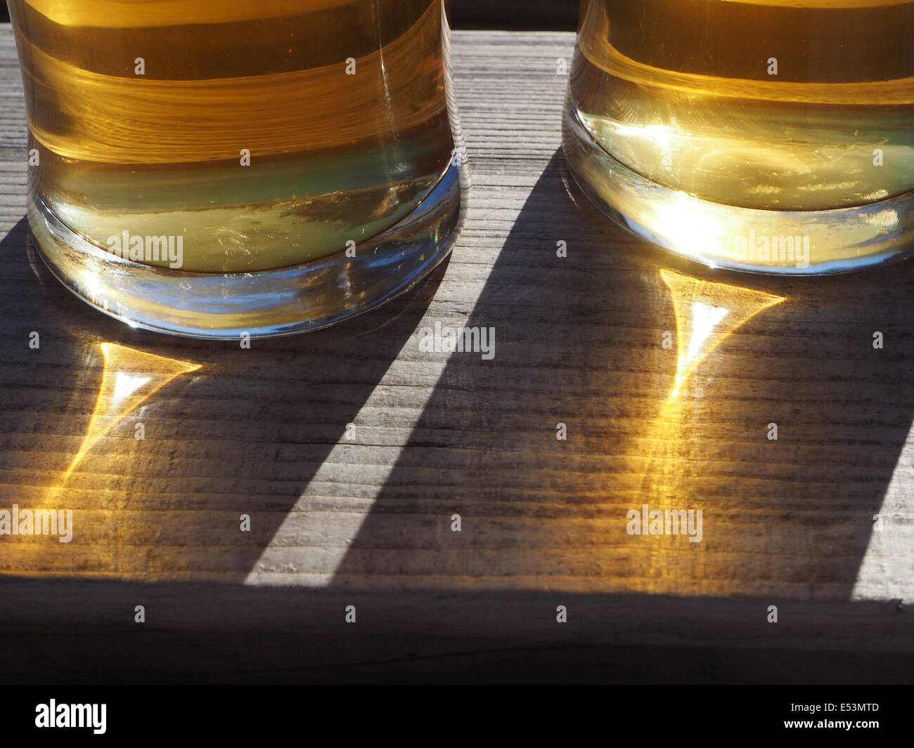 Refracted light through the base of two beer glasses creating triangular highlights in shadows on wood grain with hints of blue Stock Photo