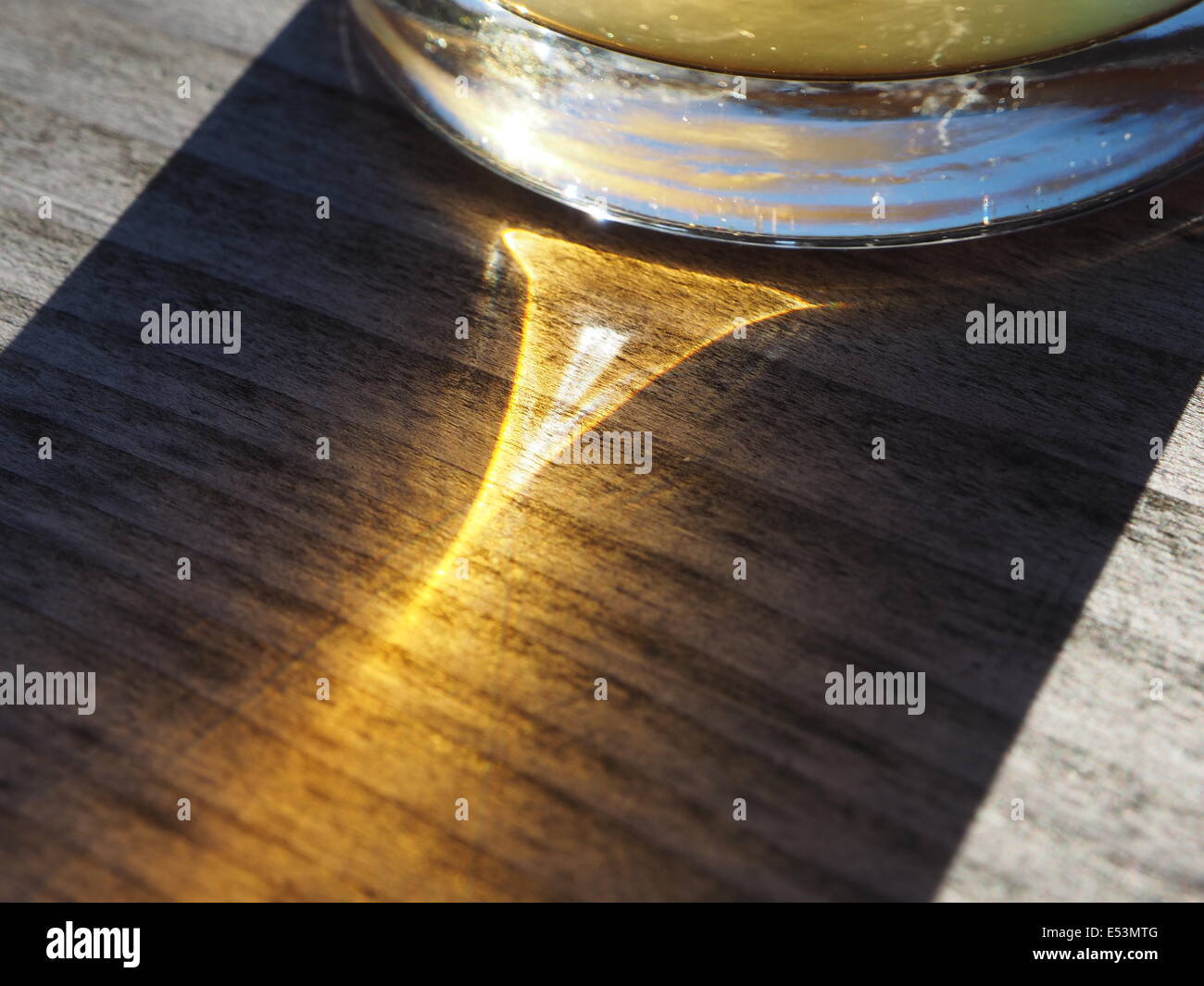 Refracted light through beer glass creating triangular highlight in the shadow on wood grain with reflection of blue sky in base Stock Photo