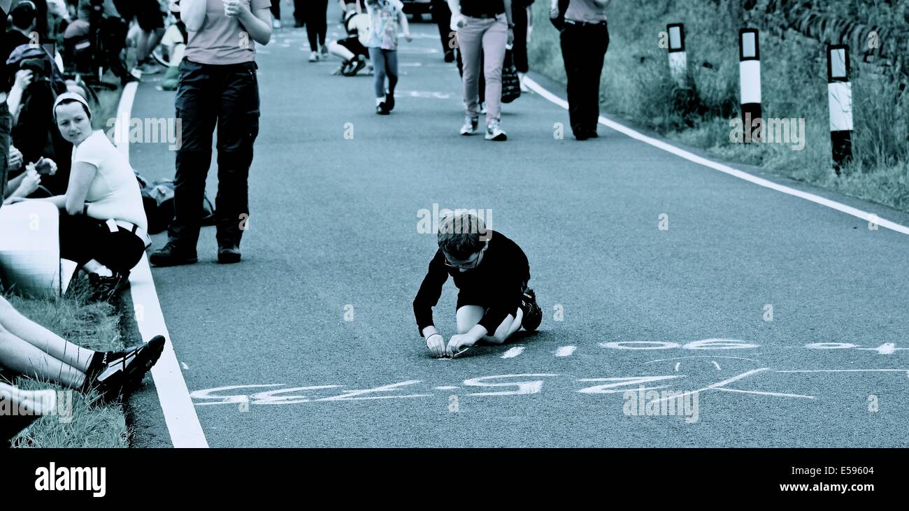 Young boy using a stone to write a message on the road on route of 2014 Tour de France South Yorkshire England Europe Stock Photo