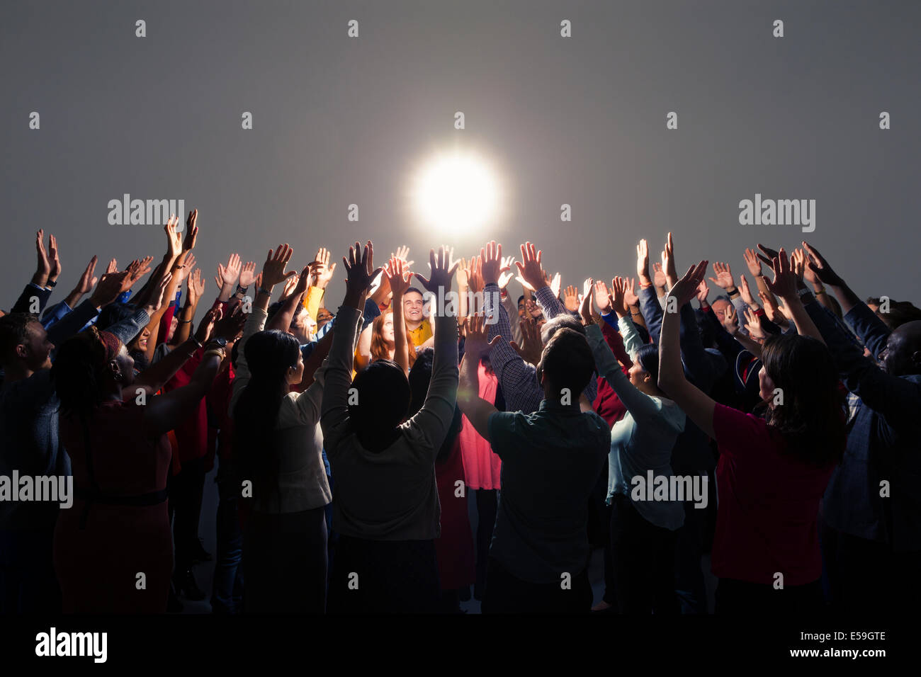 Diverse crowd with arms raised around bright light Stock Photo