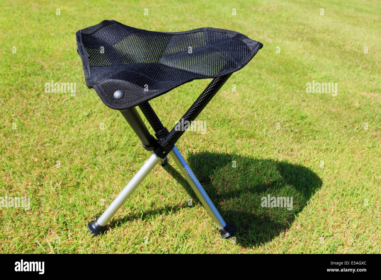 Folding portable three-legged tripod camping stool with a triangular mesh seat on grass in sunshine Stock Photo