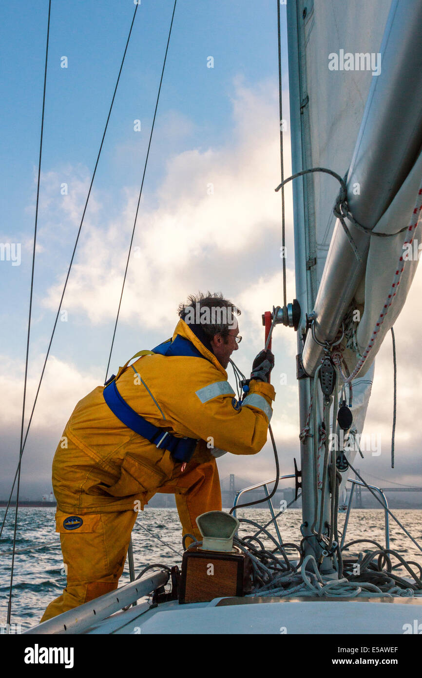 Sailor in yellow foul weather gear securing the halyard after raising the mainsail heading into cloudy weather in his sailboat Stock Photo