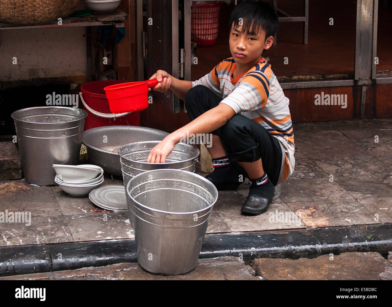 Child Labor - Sad looking young boy rinsing plates in metal buckets. Stock Photo