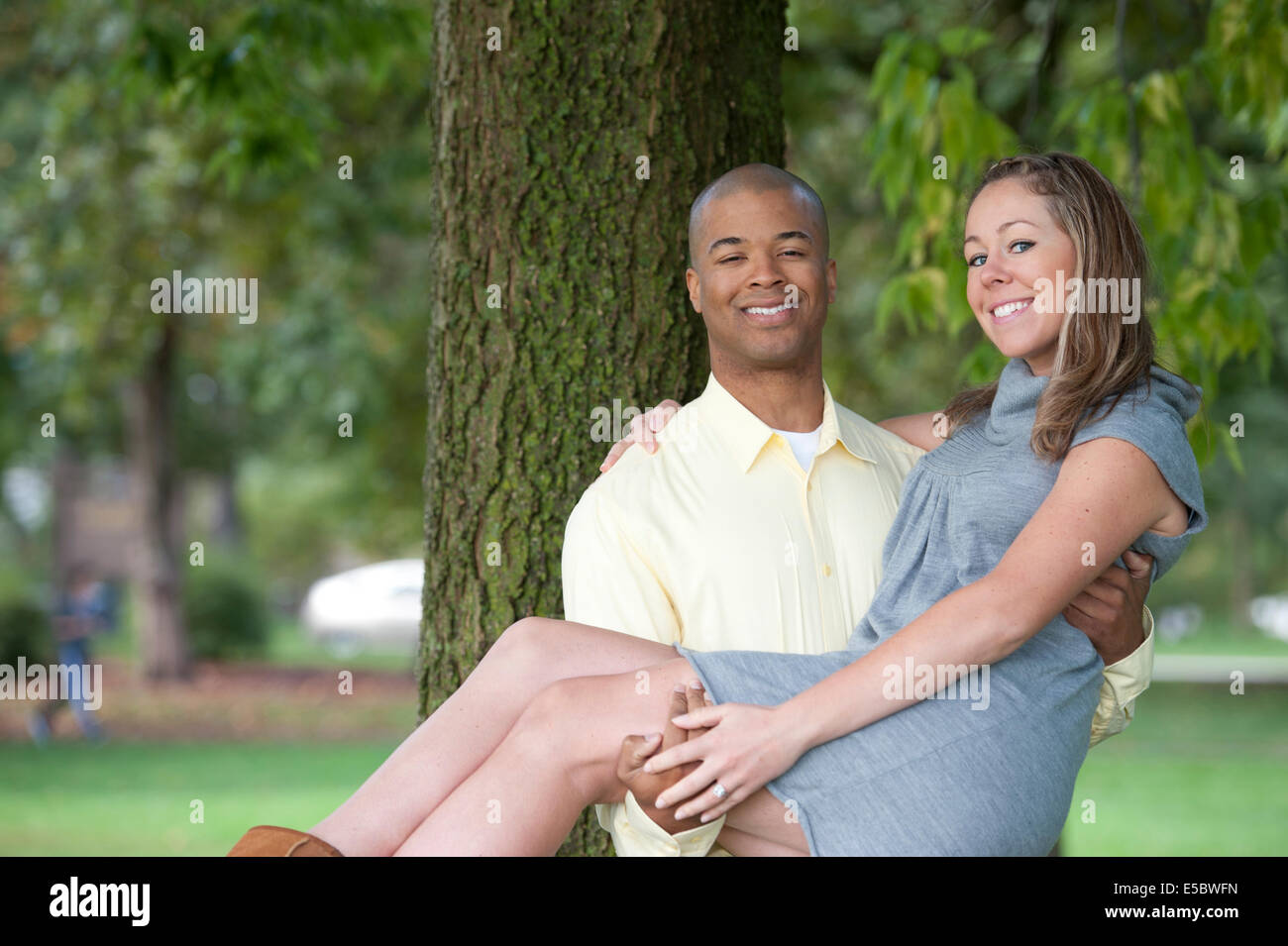 A beautiful, happy and young interracial couple posing next to a tree on a sunny day. Stock Photo
