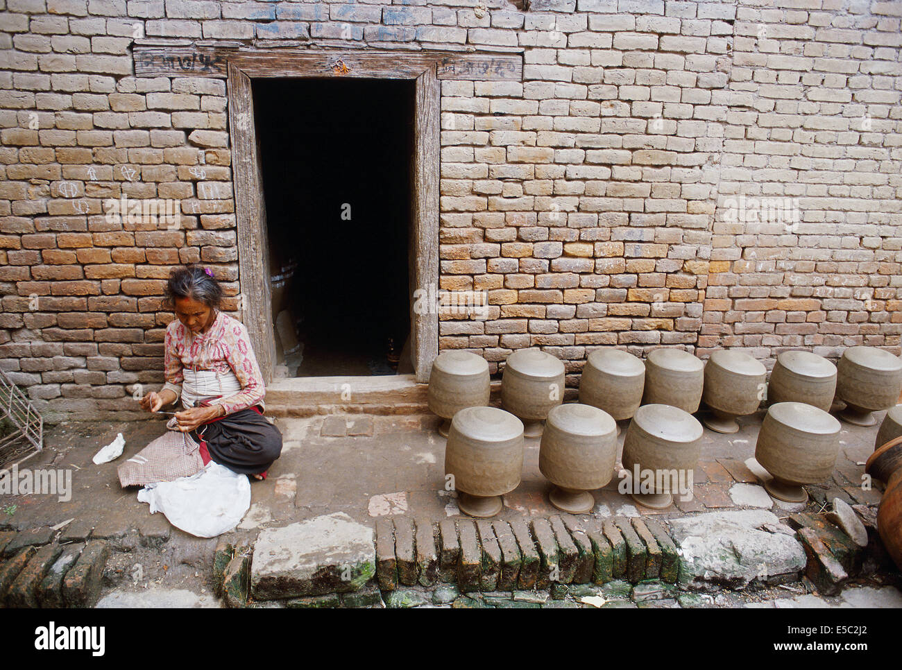 Old woman sewing in front of her house. On the right, earthen pots are drying. The woman belongs to the potter caste ( Nepal) Stock Photo