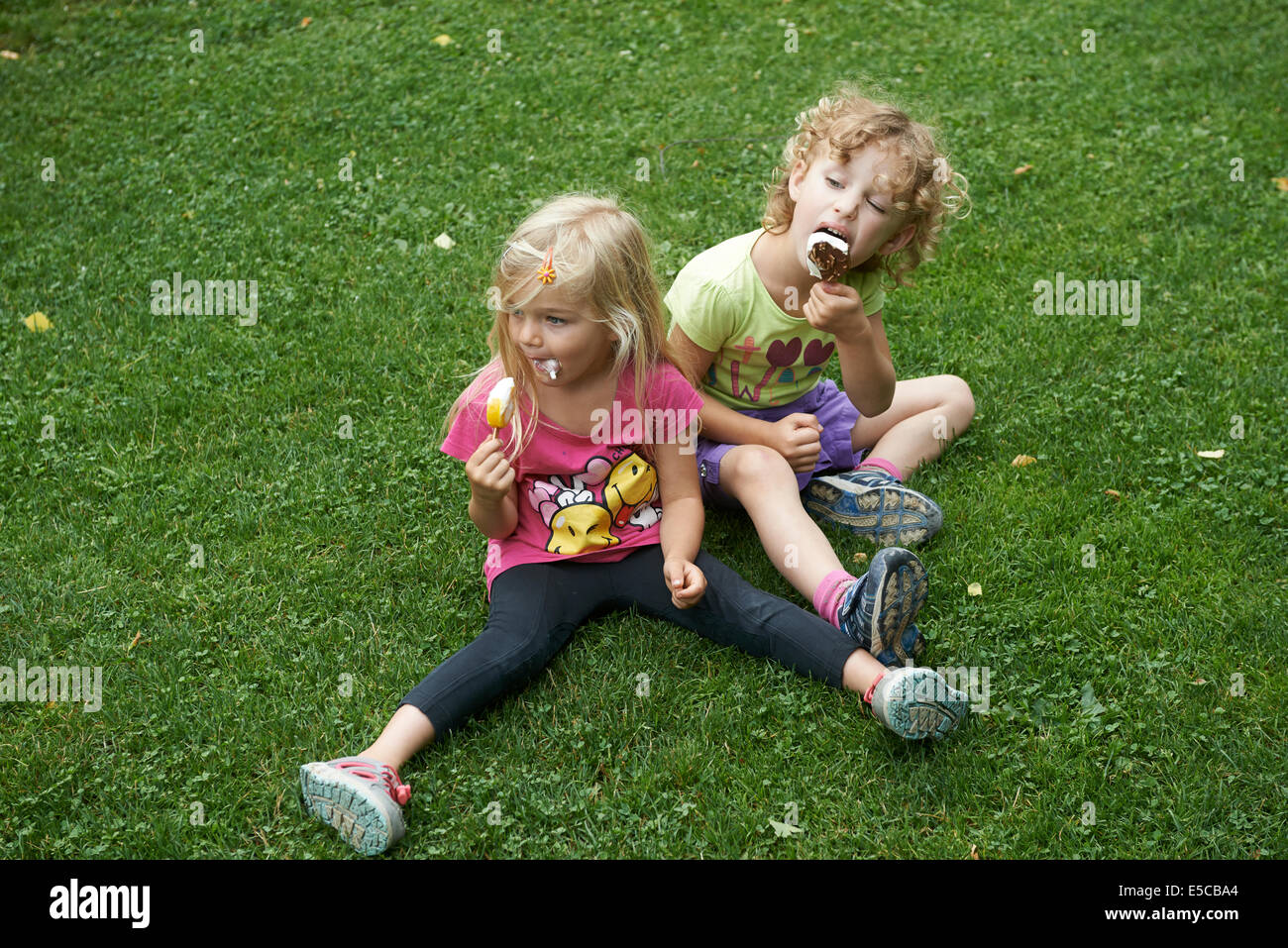 Children blond girls eating ice cream cones sitting on grass lawn, summertime Stock Photo