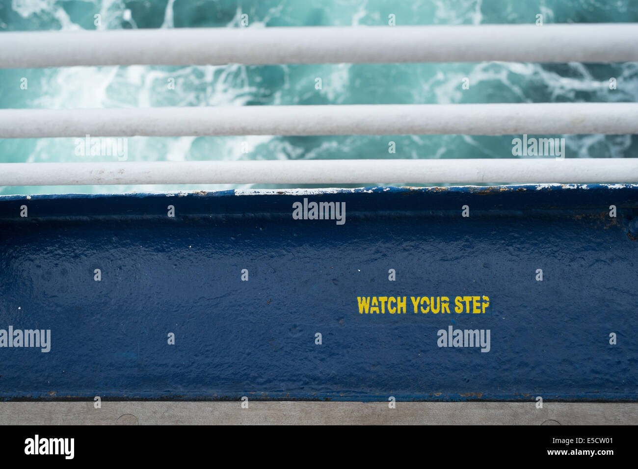 Yellow painted stencil lettering 'watch you step' on bright blue steel deck, Marco Polo cruise ship Stock Photo