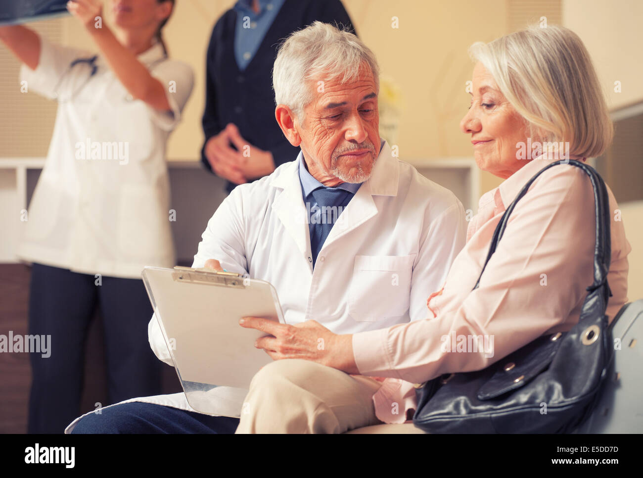 Doctors and patients in hospital waiting room. Senior male doctor explaining medical exams to woman patient. Stock Photo