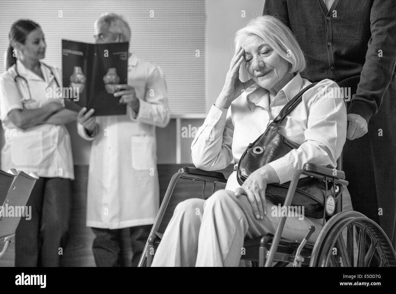 Mature woman on the wheelchair in hospital worried with her husband. Doctors analyzing scan in background. Stock Photo