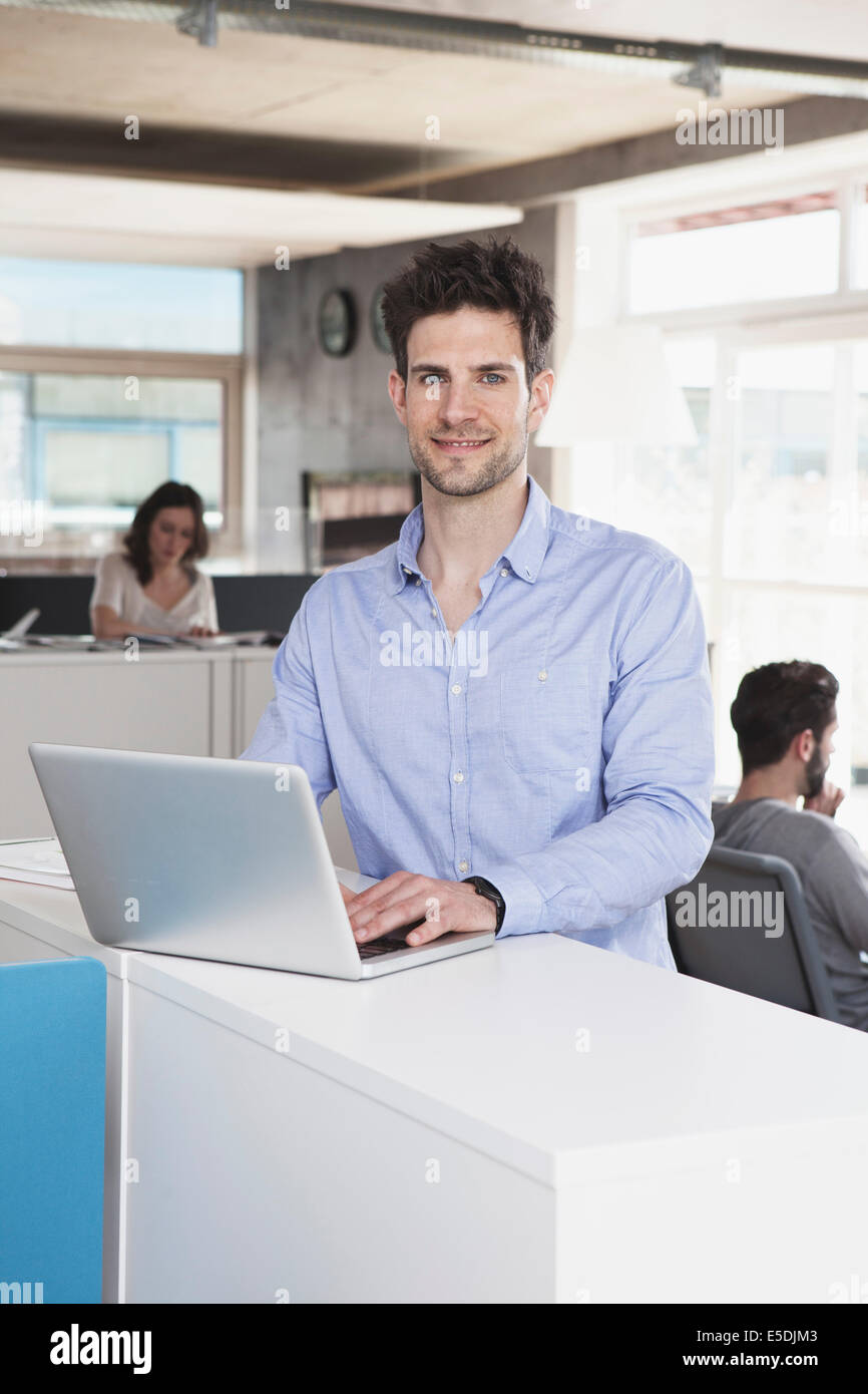 Portrait of smiling man with laptop standing in the open space office ...