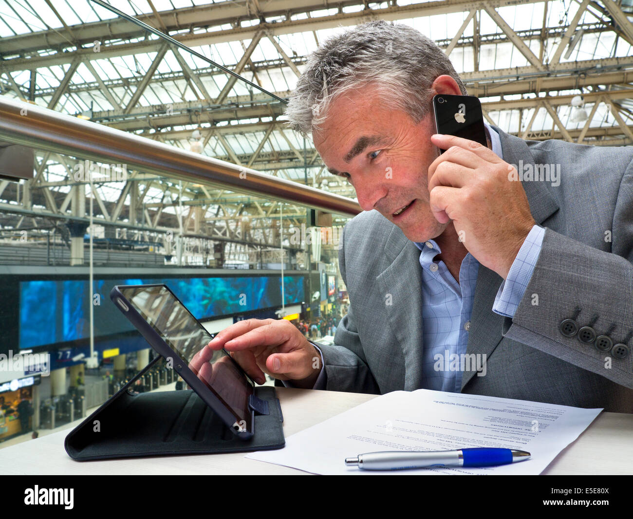 Busy mature businessman seated at cafe table on railway concourse looking at his tablet computer screen and using his iPhone Stock Photo
