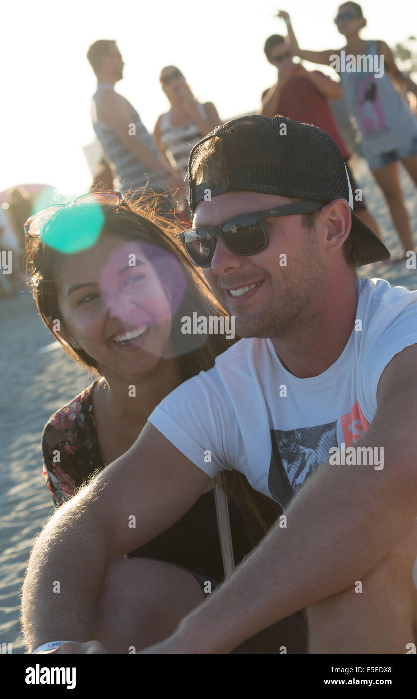 A smiling couple sitting on the beach at a music festival with lens flare across their faces and people in the background. Stock Photo