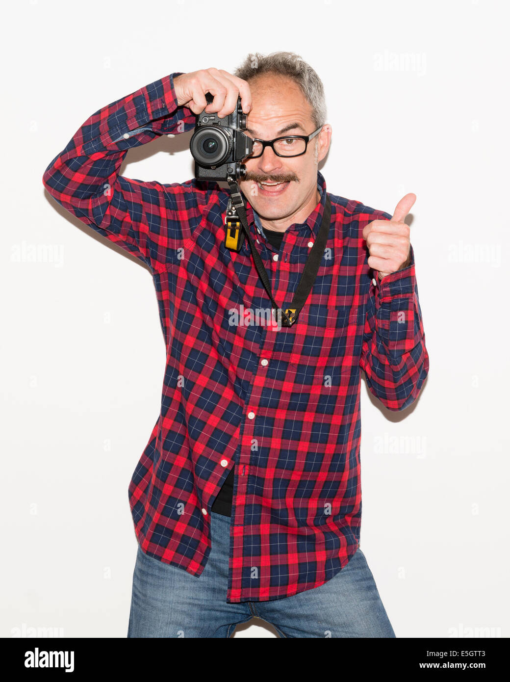 Fashion photographer guy with moustache wearing checked shirt holding a camera and taking a picture while giving thumbs up. Stock Photo