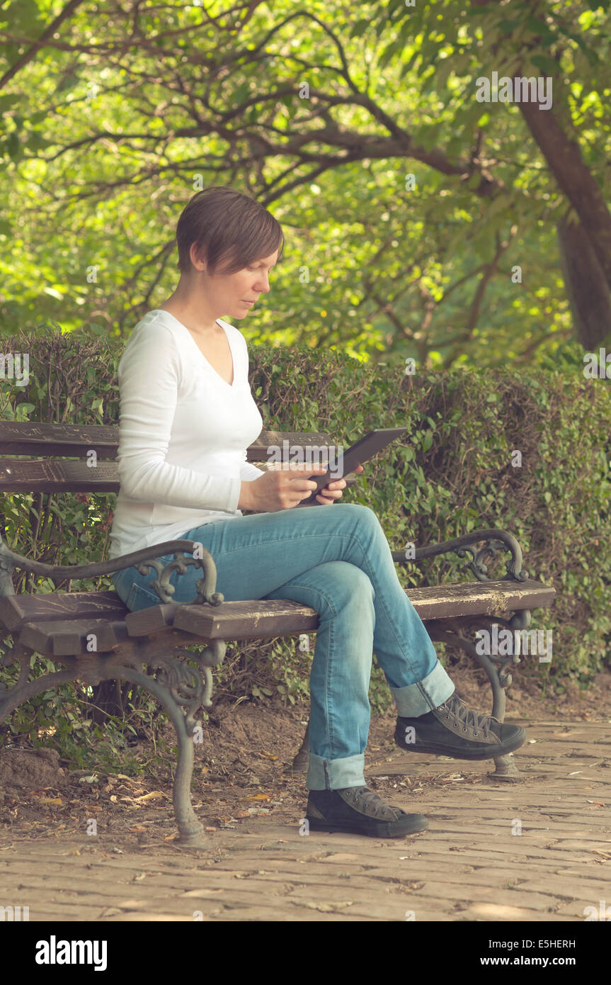 Woman using digital tablet computer and browsing the internet while sitting on a wooden bench in the park. Stock Photo