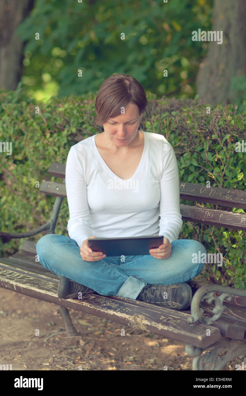 Woman using digital tablet computer and browsing the internet while sitting on a wooden bench in the park. Stock Photo