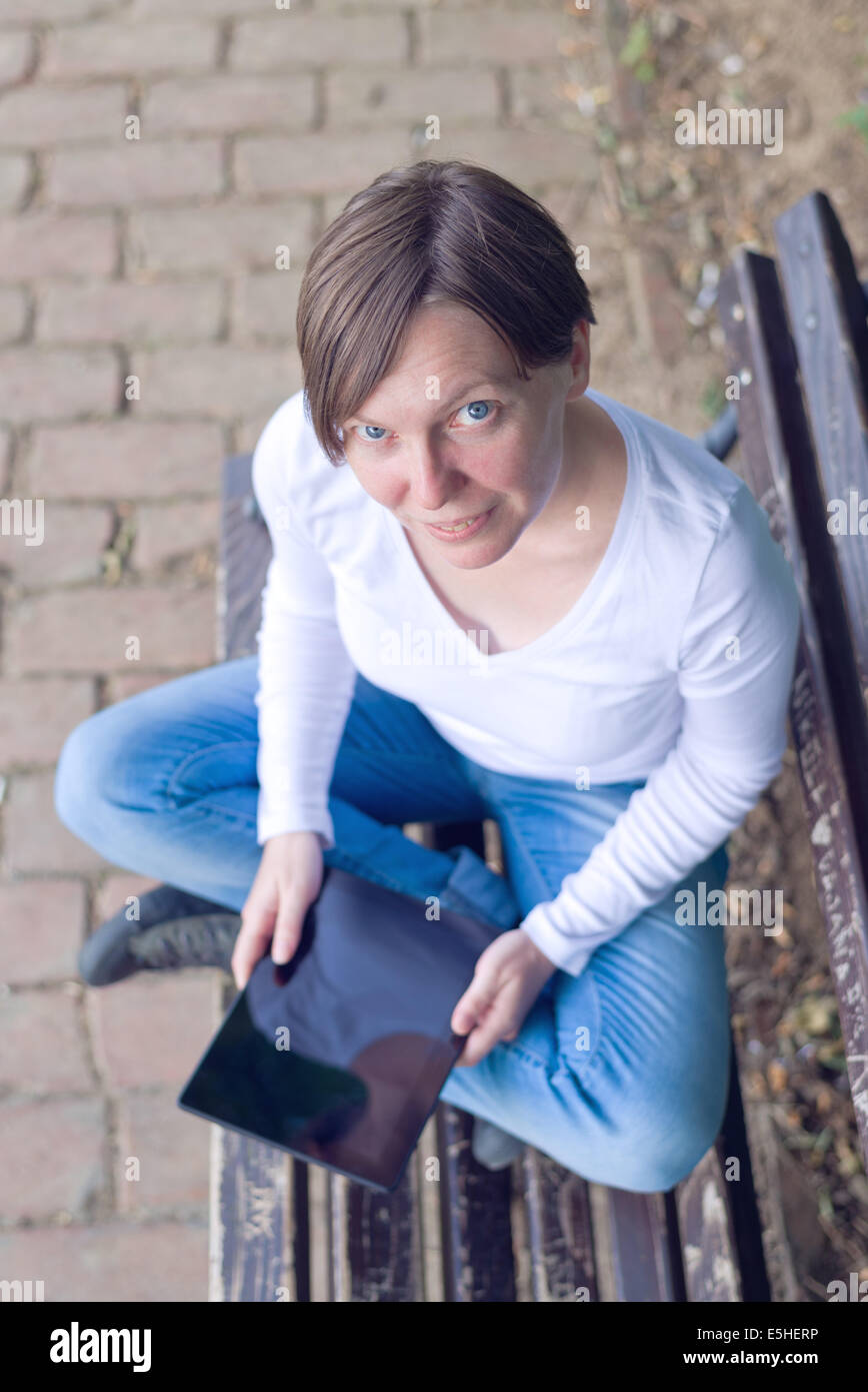 Woman holding digital tablet computer while sitting on a wooden bench in the park. Stock Photo