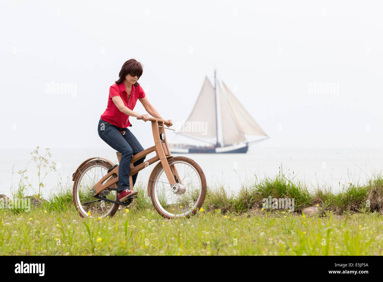 Netherlands, Venhuizen, Woman riding Bough Bike. The Dutch Design wooden bike is a creation of Jan Gunneweg. Sailing boat Stock Photo
