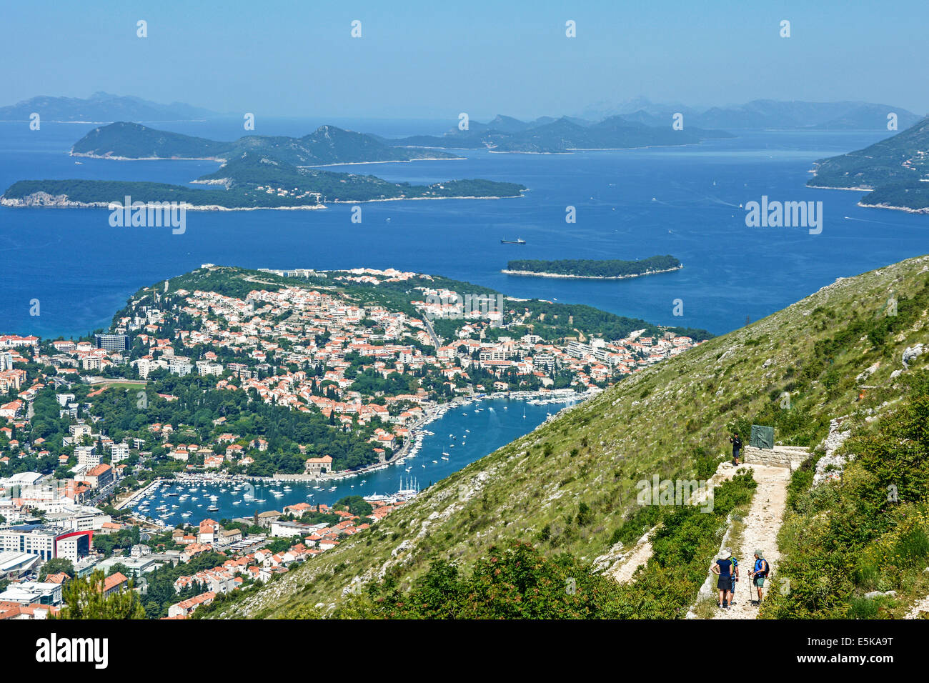 Two walkers on path leading down from Mount Srd to walled City of Dubrovnik with views towards Gruz port and islands beyond Stock Photo