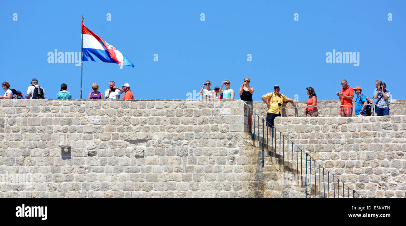 Tourists walking old city walls of Dubrovnik with Croatian national flag fluttering in breeze on hot summer blue sky day Croatia Dalmatia Adriatic Stock Photo