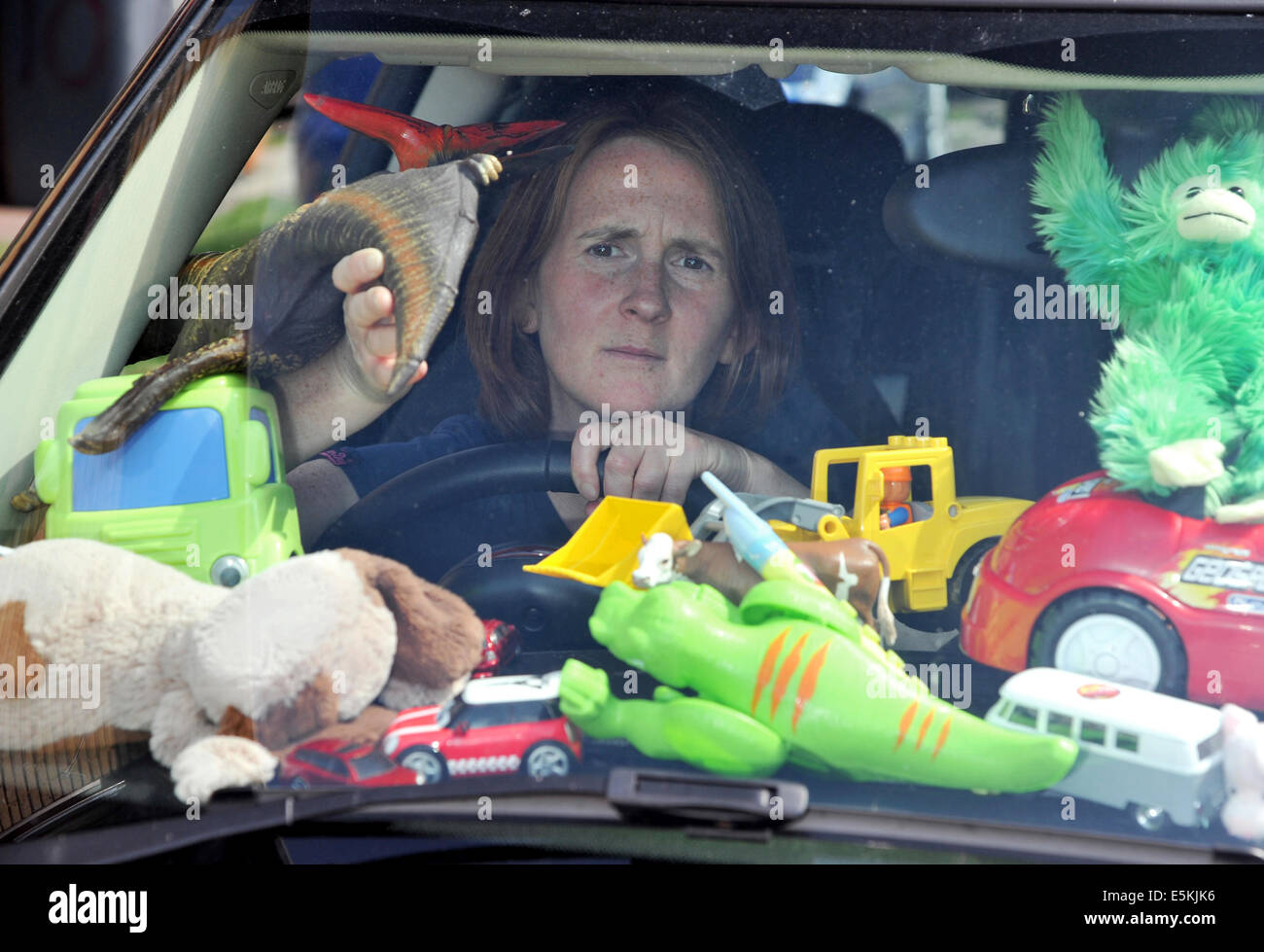 Stressed mother overwhelmed by piles of kids toys in her car, peeps through a gap to see out the windscreen Stock Photo