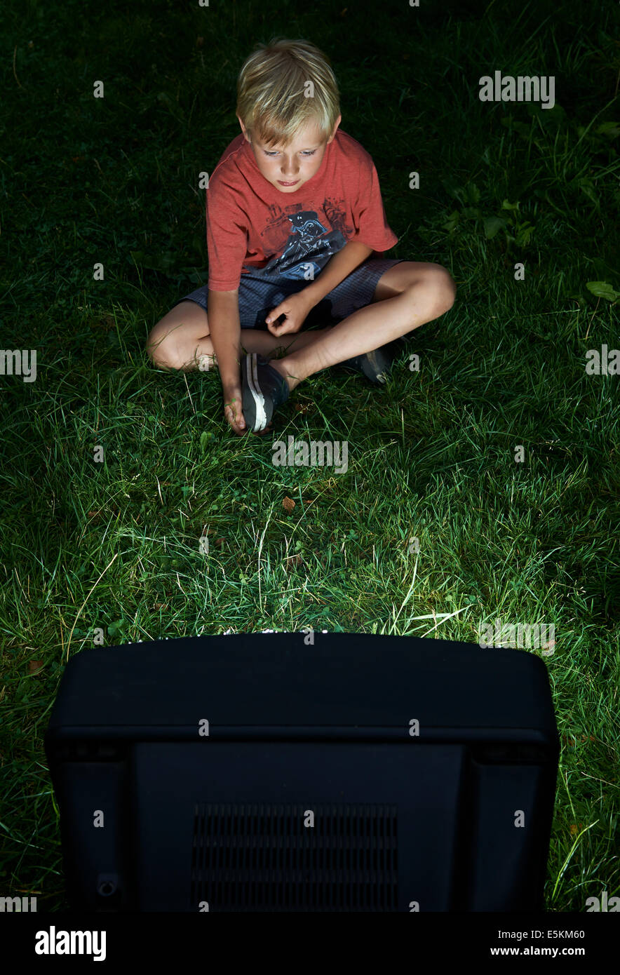 Child Blond Boy Watching Television in Yard, outside in green grass lawn, emotional, facial expression, grimace, grinning Stock Photo