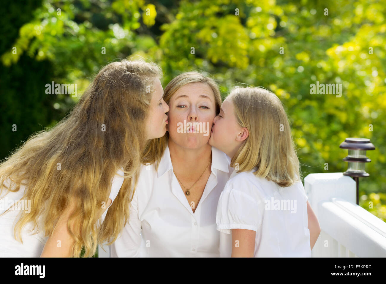 Front view of mother, displaying fish lips, being kissed by her two daughters outdoors on patio with woods in background Stock Photo