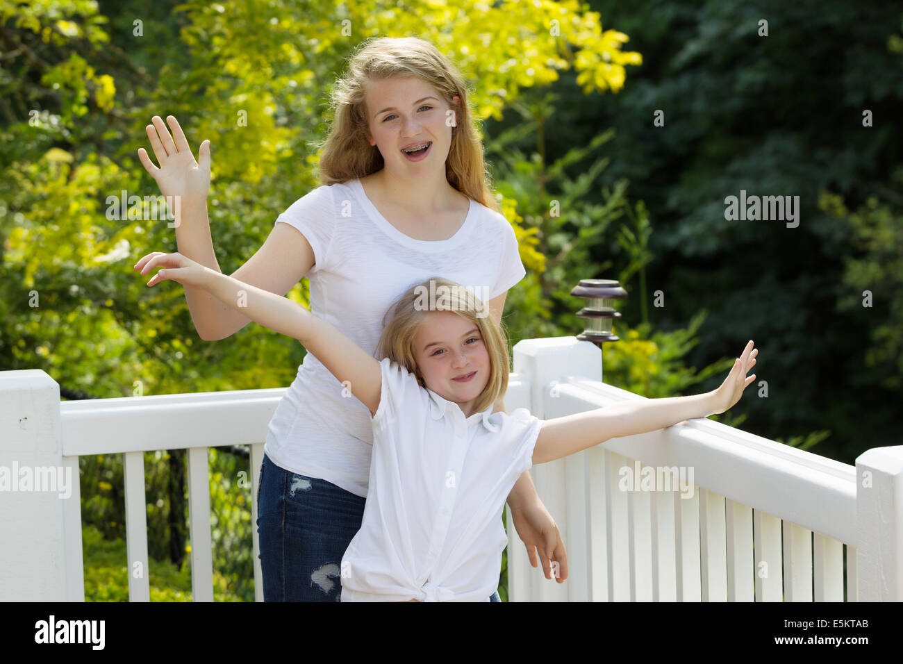 Front view, looking forward, of two sisters waving their arms while outdoors on patio with blurred out trees in background Stock Photo