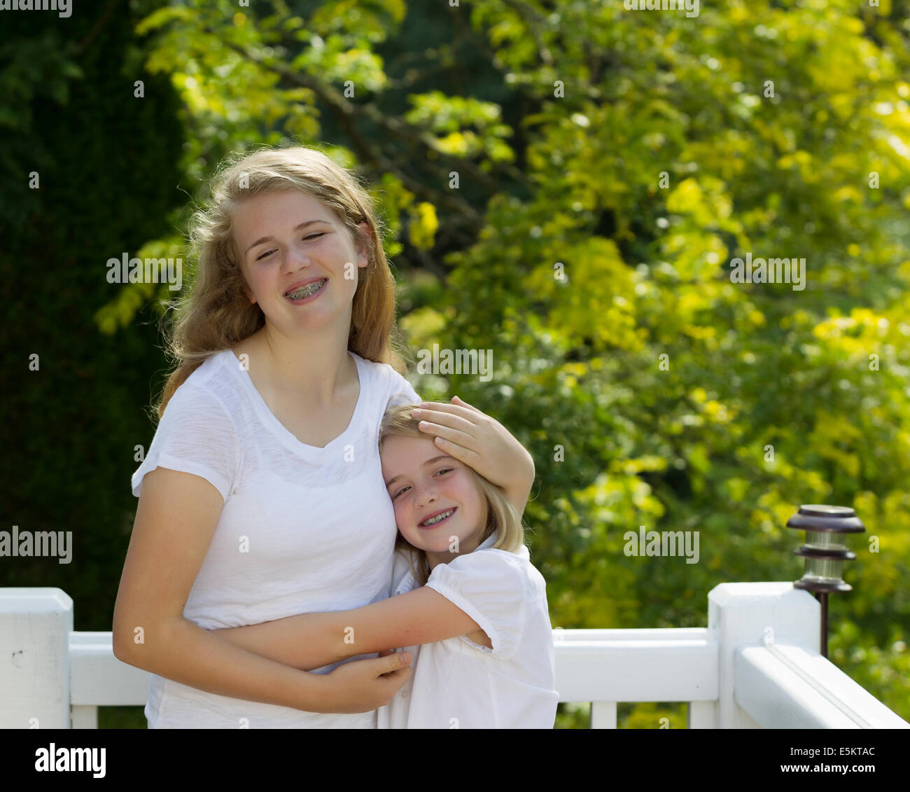 Front view of two sisters hugging each other while outdoors on patio with blurred out trees in background Stock Photo