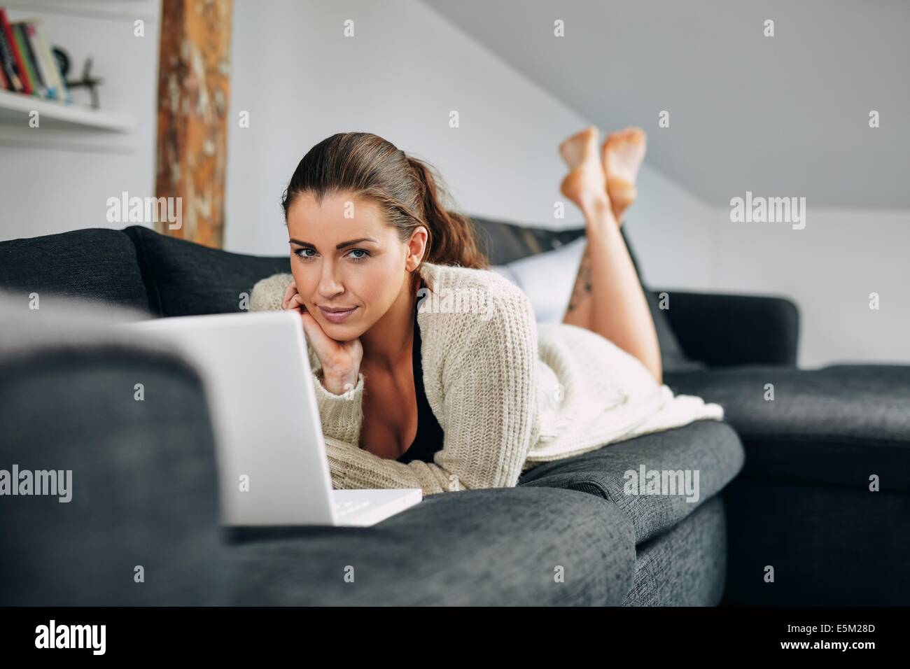 Portrait of pretty young female lying on sofa with a laptop looking at ...