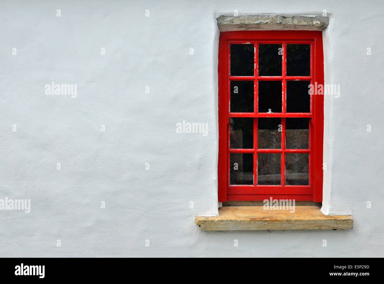 Small red painted sash window with stone cill and lintel set in the white-painted wall of a traditional Irish cottage. Stock Photo